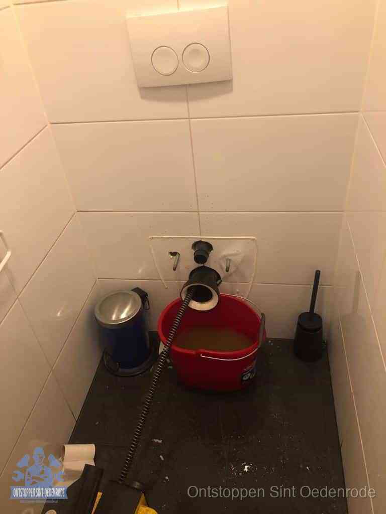 Een ontstopte wand-wc met verwijderde toiletpot waar een ontstoppingsveer in de wanduitgang verdwijnt, het achterhalen waar de verstopping zit in Sint Oedenrode