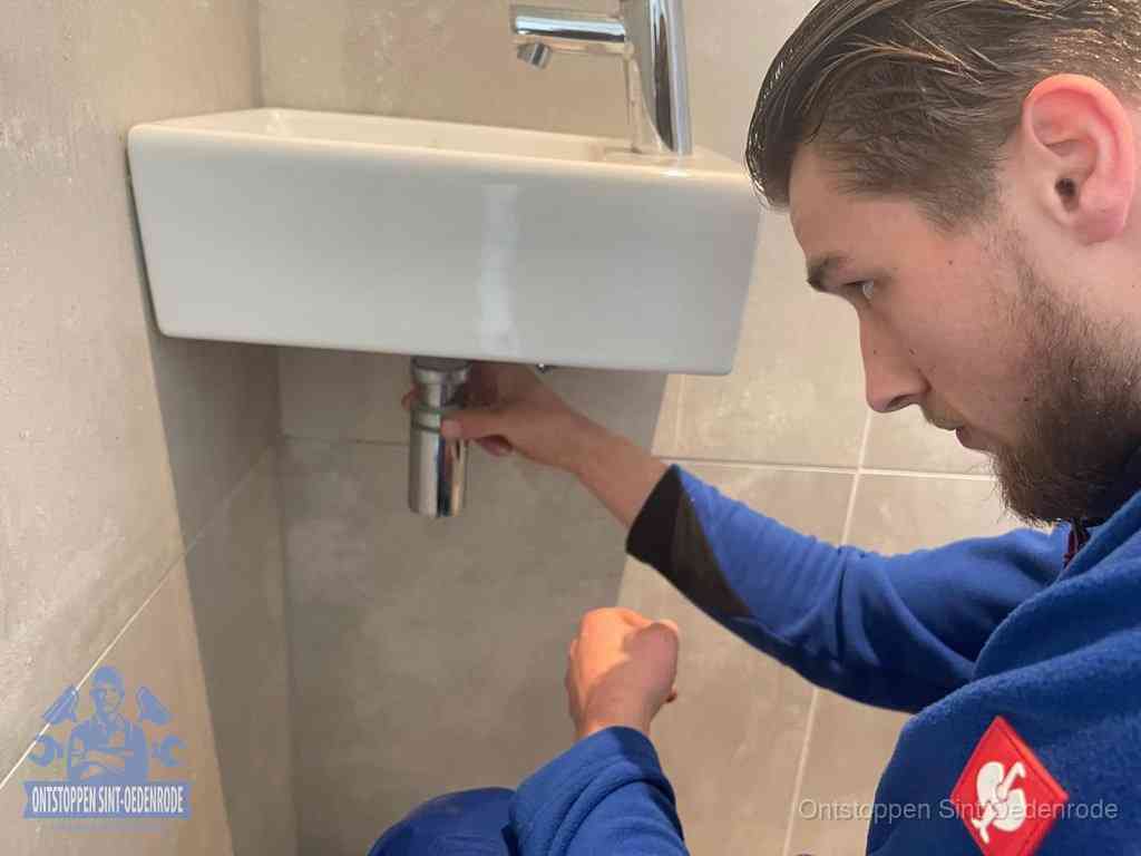 Een monteur draait met de hand de zwanenhalssifon onder een fonteinwasbak in de toiletruimte los om de bron van rioollucht in Sint Oedenrode te vinden