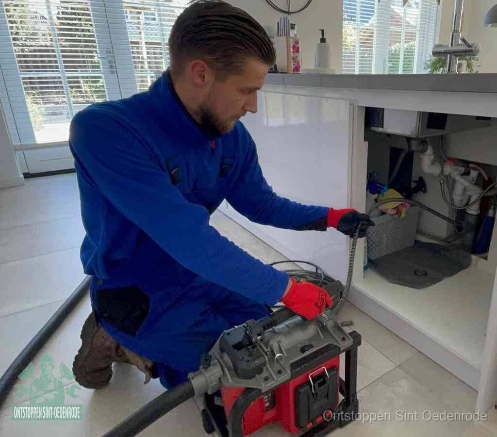 Een monteur met rode handschoenen knielt in een keuken en voert een elektrische ontstoppingsveer in tijdens het professioneel afvoer ontstoppen in Sint Oedenrode